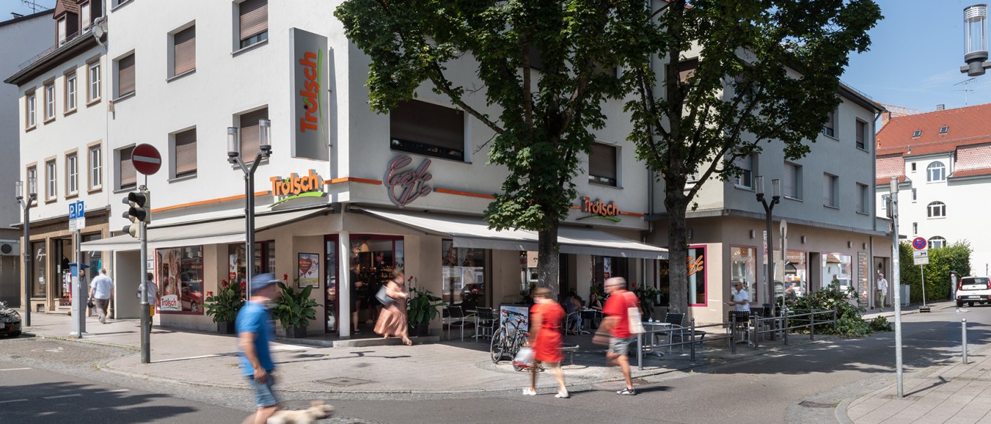 Straßenszene mit Café Trölsch an einer Ecke, Menschen und ein Hund gehen vorbei, Bäume spenden Schatten., © Trölsch GmbH