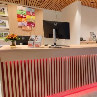 Moderner Empfangsbereich mit Holztheke, Computern, Broschüren und Blumenvasen. Helle Beleuchtung und stilvolle Einrichtung., © Larissa Philipp