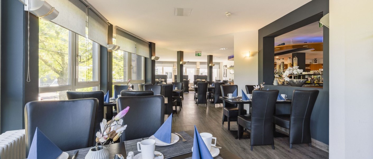 Moderner Frühstücksbereich mit gedeckten Tischen, blauen Servietten und großen Fenstern, die viel Licht hereinlassen., © Plaza INN Stuttgart-Filderstadt Moderner Frühstücksbereich mit gedeckten Tischen, blauen Servietten und großen Fenstern, die viel Licht hereinlassen., © Plaza INN Stuttgart-Filderstadt