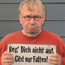 Ein Mann mit Brille und rotem Shirt h&auml;lt ein Schild mit der Aufschrift: 'Reg' Dich nicht auf. Gibt nur Falten!' vor einem blauen Hintergrund., &copy; links im Bild