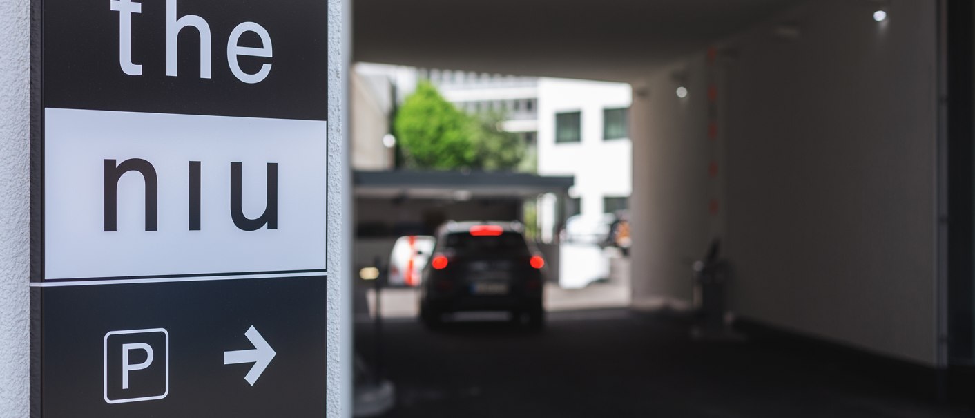 Schild mit 'the niu' und Parkplatzsymbol zeigt nach rechts. Auto fährt in Tiefgarage., © the niu Form - Novum Hospitality