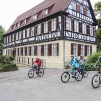 Vier Radfahrer fahren an einem historischen Fachwerkhaus in Kirchheim unter Teck vorbei. Das Gebäude hat weiße Wände und braune Fensterläden., © Schwäbische Alb Tourismusverband e.V.