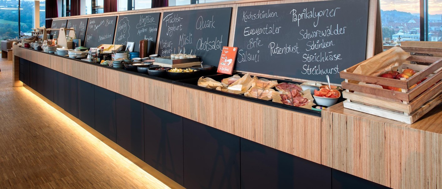 Ein reichhaltiges Frühstücksbuffet mit verschiedenen Speisen und Tafeln, die das Angebot beschreiben. Im Hintergrund sind Fenster mit Ausblick., © Hotel am Remspark Ein reichhaltiges Frühstücksbuffet mit verschiedenen Speisen und Tafeln, die das Angebot beschreiben. Im Hintergrund sind Fenster mit Ausblick., © Hotel am Remspark