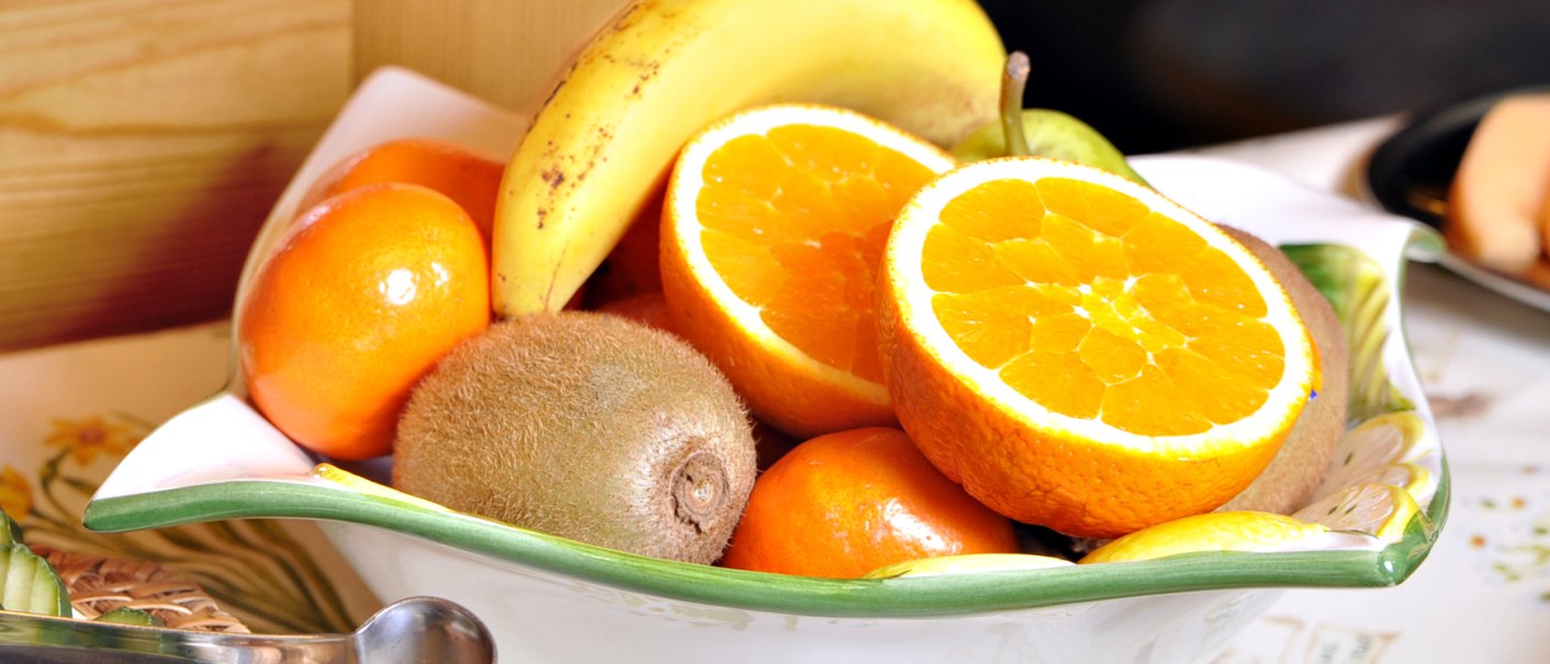 Eine Schale mit frischen Früchten: Orangen, Banane, Kiwi und Mandarinen. Im Vordergrund liegt eine Zange auf einem Tisch., © TOMAS