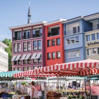 Wochenmarkt in Stuttgart mit bunten Marktst&auml;nden und Geb&auml;uden im Hintergrund. Menschen schlendern zwischen den St&auml;nden, w&auml;hrend die Sonne scheint., &copy; SMG, trickytine