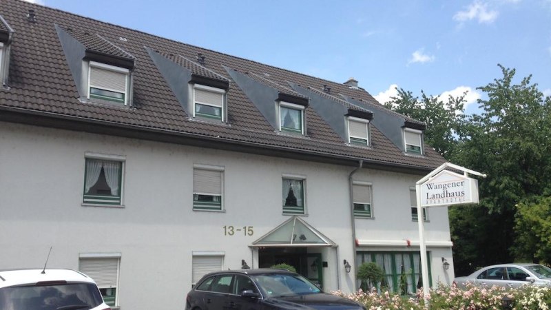Das Aparthotel Wangener Landhaus mit grauem Dach und wei&szlig;en W&auml;nden. Vor dem Geb&auml;ude sind Autos geparkt. Ein Schild zeigt den Hotelnamen., &copy; Aparthotel Wangener Landhaus