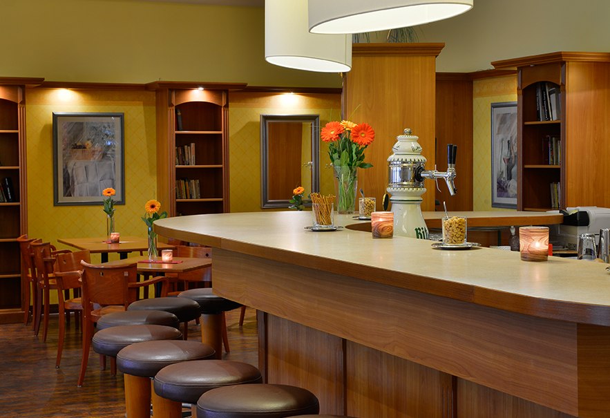 Eine elegante Bar mit Holztheke, Barhockern und stilvoller Beleuchtung. Blumen und Kerzen dekorieren die Theke, im Hintergrund sind Tische und Regale., &copy; Amber Hotel Leonberg / Stuttgart