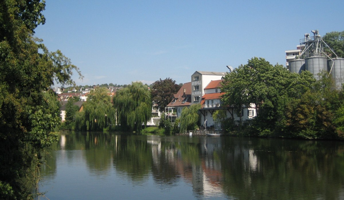 Ein ruhiger Fluss mit Bäumen und Gebäuden am Ufer, darunter eine Mühle mit der Aufschrift 'Mühle'. Der Himmel ist klar und blau., © Land der 1000 Hügel - Kraichgau-Stromberg