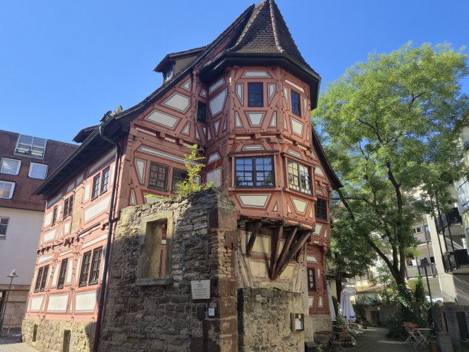 Ein historisches Fachwerkhaus mit roten Balken und steilem Dach in einer Altstadt. Ein Baum und blauer Himmel im Hintergrund., © Cool-Tours StattReisen Ein historisches Fachwerkhaus mit roten Balken und steilem Dach in einer Altstadt. Ein Baum und blauer Himmel im Hintergrund., © Cool-Tours StattReisen