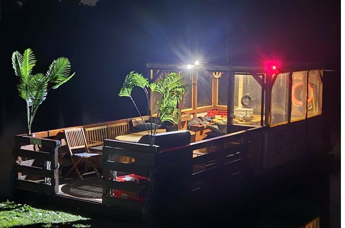Ein beleuchtetes Floß mit Pflanzen und Möbeln auf einem dunklen Gewässer bei Nacht., © Cool-Tours StattReisen