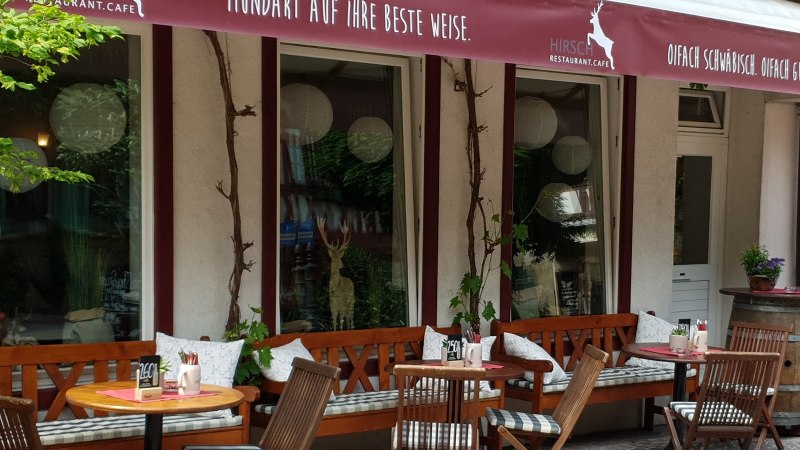 Gemütlicher Außenbereich des Restaurant Café Hirsch mit Holzbänken, Tischen und Pflanzen. Ein rotes Vordach mit Schriftzug ist sichtbar., © Restaurant Café Hirsch