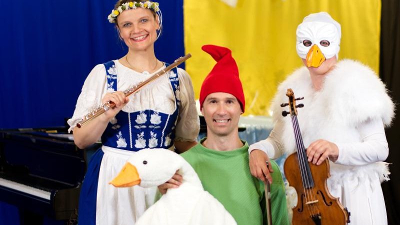 Drei Personen in bunten Kostümen, eine mit Flöte, eine mit Geige und eine mit Gans-Puppe. Hintergrund in Blau und Gelb., © Backnang