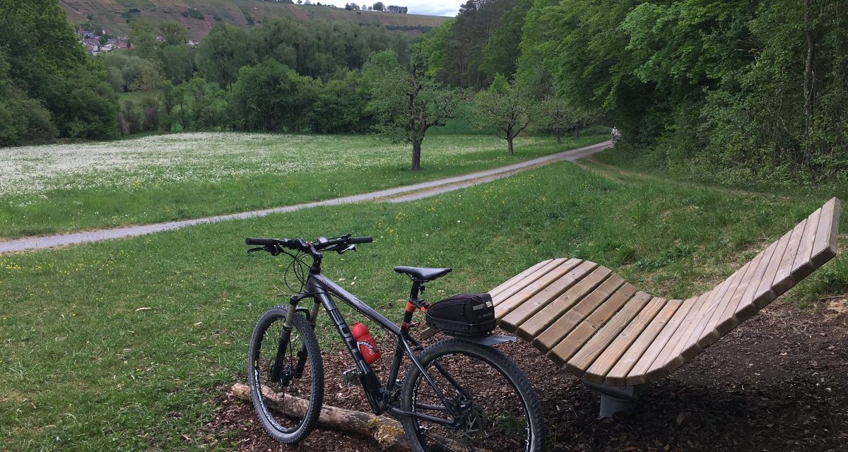 Fahrrad neben geschwungener Holzbank auf Wiese, umgeben von Bäumen und Hügeln im Hintergrund., © Land der 1000 Hügel - Kraichgau-Stromberg Fahrrad neben geschwungener Holzbank auf Wiese, umgeben von Bäumen und Hügeln im Hintergrund., © Land der 1000 Hügel - Kraichgau-Stromberg