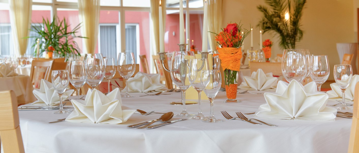Eleganter Tisch im Hotel-Restaurant mit weißen Servietten, Gläsern und Kerzen. Blumendekoration in Orange. Große Fenster im Hintergrund., © Hotel-Restaurant "Talblick"