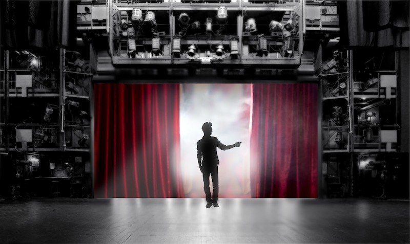 Ein Mann steht auf einer Bühne vor einem roten Vorhang, umgeben von Bühnentechnik. Der Hintergrund ist schwarz-weiß, der Vorhang rot., © LIMA-Theater