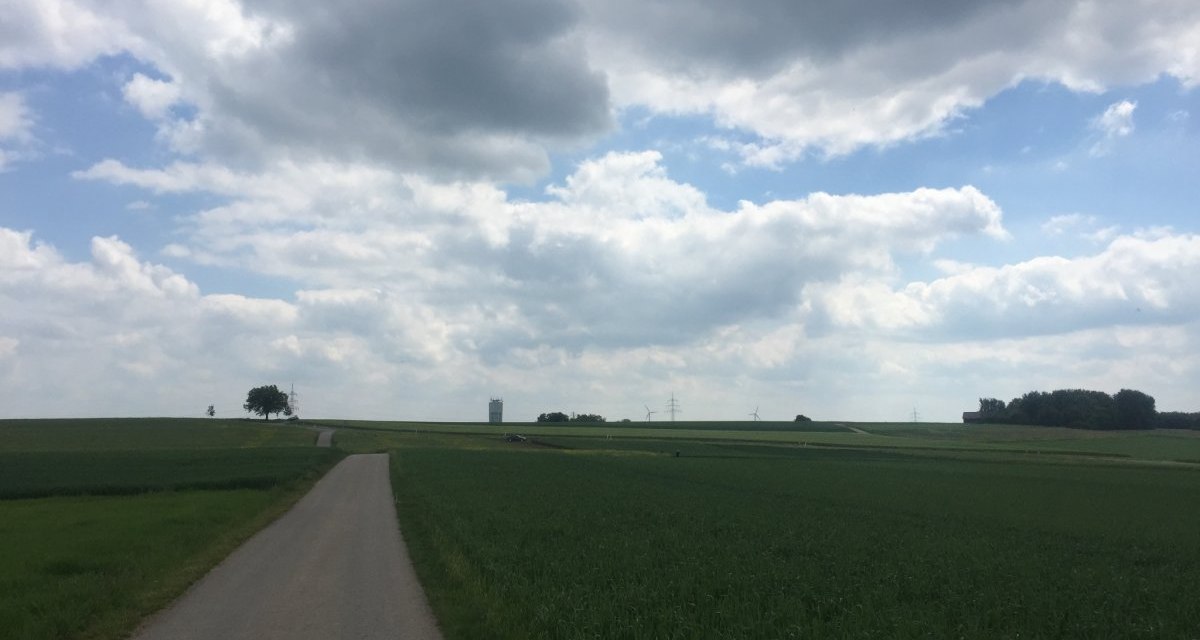 Ein schmaler Weg führt durch grüne Felder unter einem bewölkten Himmel. In der Ferne ist ein Wasserturm zu sehen, umgeben von Windrädern und Bäumen., © www.pro-cycl.de Ein schmaler Weg führt durch grüne Felder unter einem bewölkten Himmel. In der Ferne ist ein Wasserturm zu sehen, umgeben von Windrädern und Bäumen., © www.pro-cycl.de