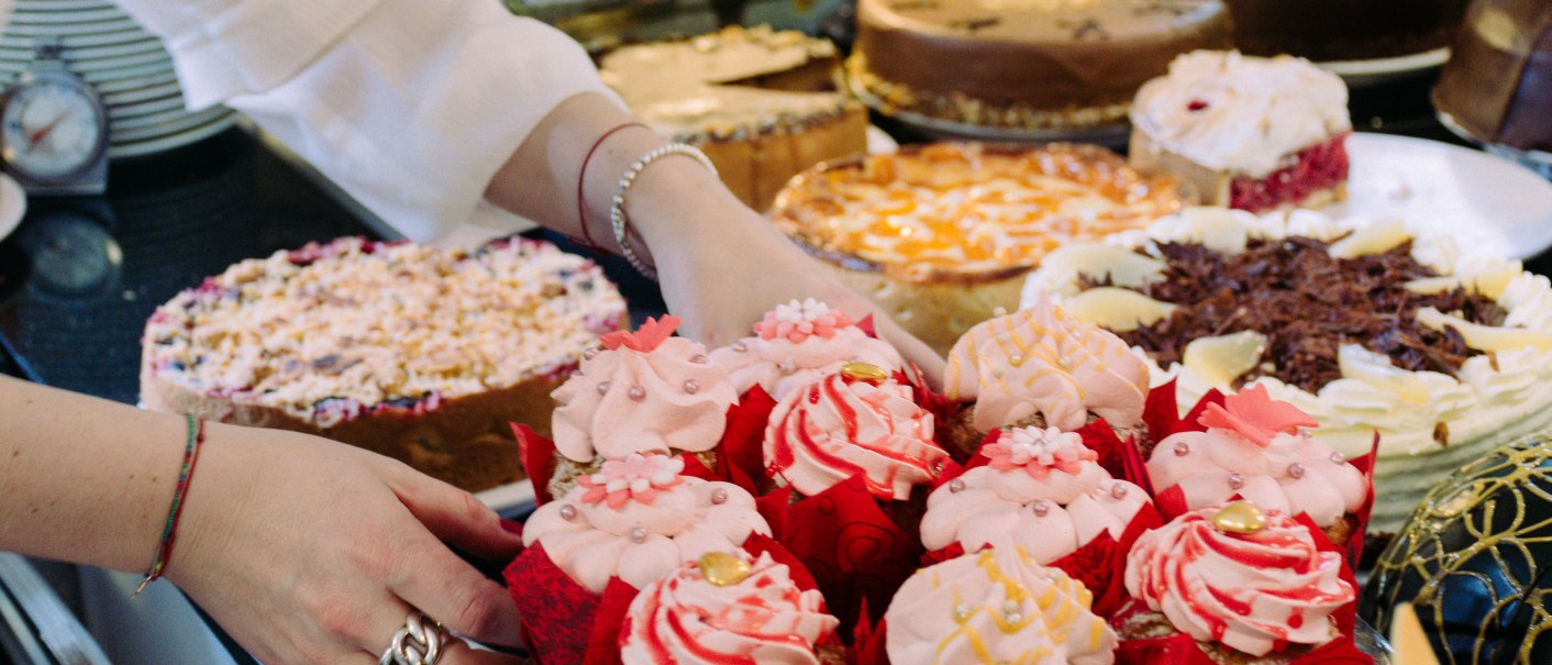 Eine Person arrangiert rosa Cupcakes auf einem Tisch mit verschiedenen Kuchen und Torten. Die Cupcakes sind mit rosa Zuckerguss und Dekorationen verziert., &copy; Sch&uuml;mli