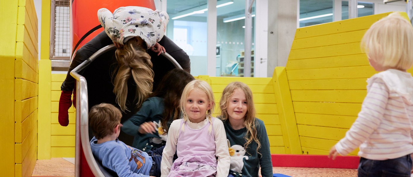 Kinder spielen auf einer Rutsche in einem Indoor-Spielplatz. Die Umgebung ist bunt mit gelben Wänden und Spielgeräten., © Julia Ochs Kinder spielen auf einer Rutsche in einem Indoor-Spielplatz. Die Umgebung ist bunt mit gelben Wänden und Spielgeräten., © Julia Ochs