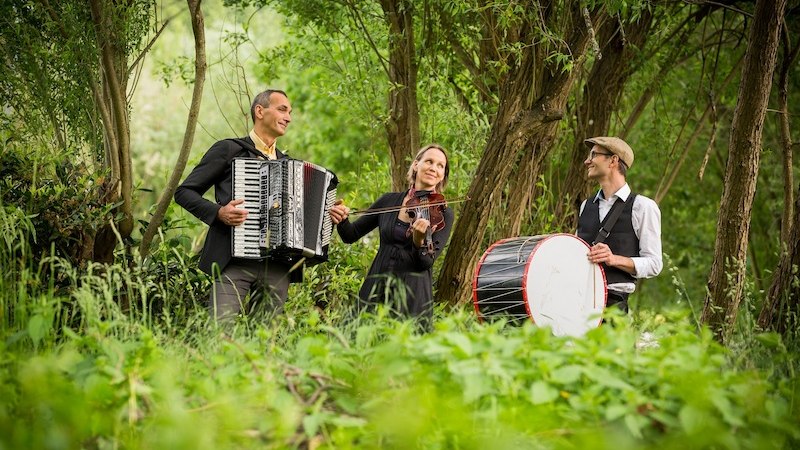 Drei Musiker mit Akkordeon, Geige und Trommel spielen in einem grünen Waldstück. Sie wirken fröhlich und entspannt., © Kulturhaus Schwanen Drei Musiker mit Akkordeon, Geige und Trommel spielen in einem grünen Waldstück. Sie wirken fröhlich und entspannt., © Kulturhaus Schwanen