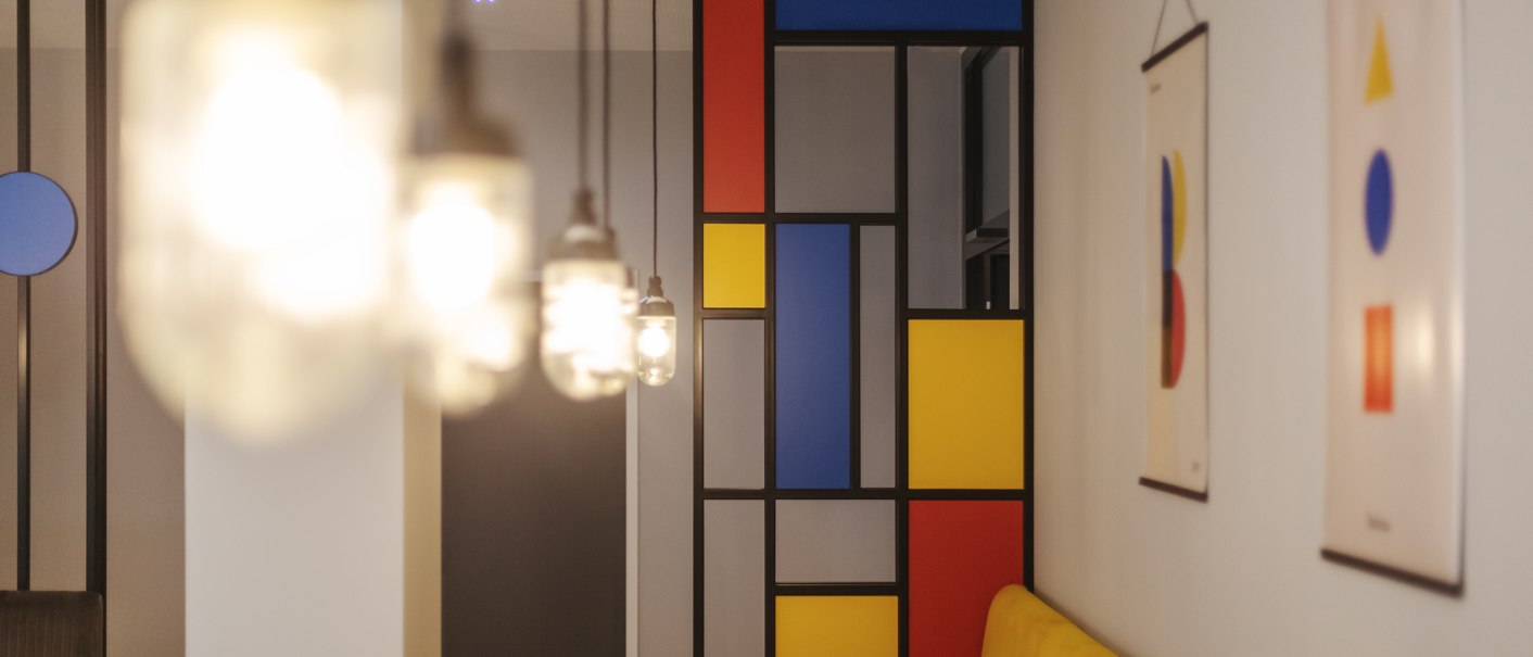 Restaurant mit geometrischem Wanddesign in Rot, Blau und Gelb. Hängende Lampen im Vordergrund, moderne Kunst an den Wänden., © the niu Form - Novum Hospitality