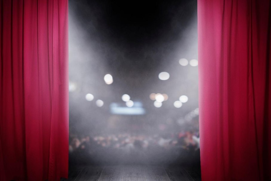 Ein roter Vorhang &ouml;ffnet sich und gibt den Blick auf eine B&uuml;hne frei. Im Hintergrund ist ein unscharfes Publikum zu sehen., &copy; AdobeStock 242383041 alphaspirit