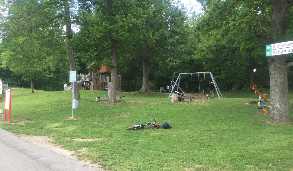 Spielplatz mit Schaukeln und Kletterger&uuml;st, umgeben von B&auml;umen. Ein Fahrrad liegt auf dem Rasen. Wegweiser zu Kleintierzoo und Reitanlage., &copy; www.pro-cycl.de