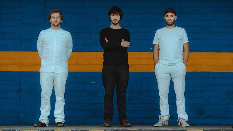 Drei Männer stehen nebeneinander vor einer blau-gelben Wand. Zwei tragen weiße Kleidung, einer schwarze. Sie blicken ernst in die Kamera., © Kulturhaus Schwanen Drei Männer stehen nebeneinander vor einer blau-gelben Wand. Zwei tragen weiße Kleidung, einer schwarze. Sie blicken ernst in die Kamera., © Kulturhaus Schwanen