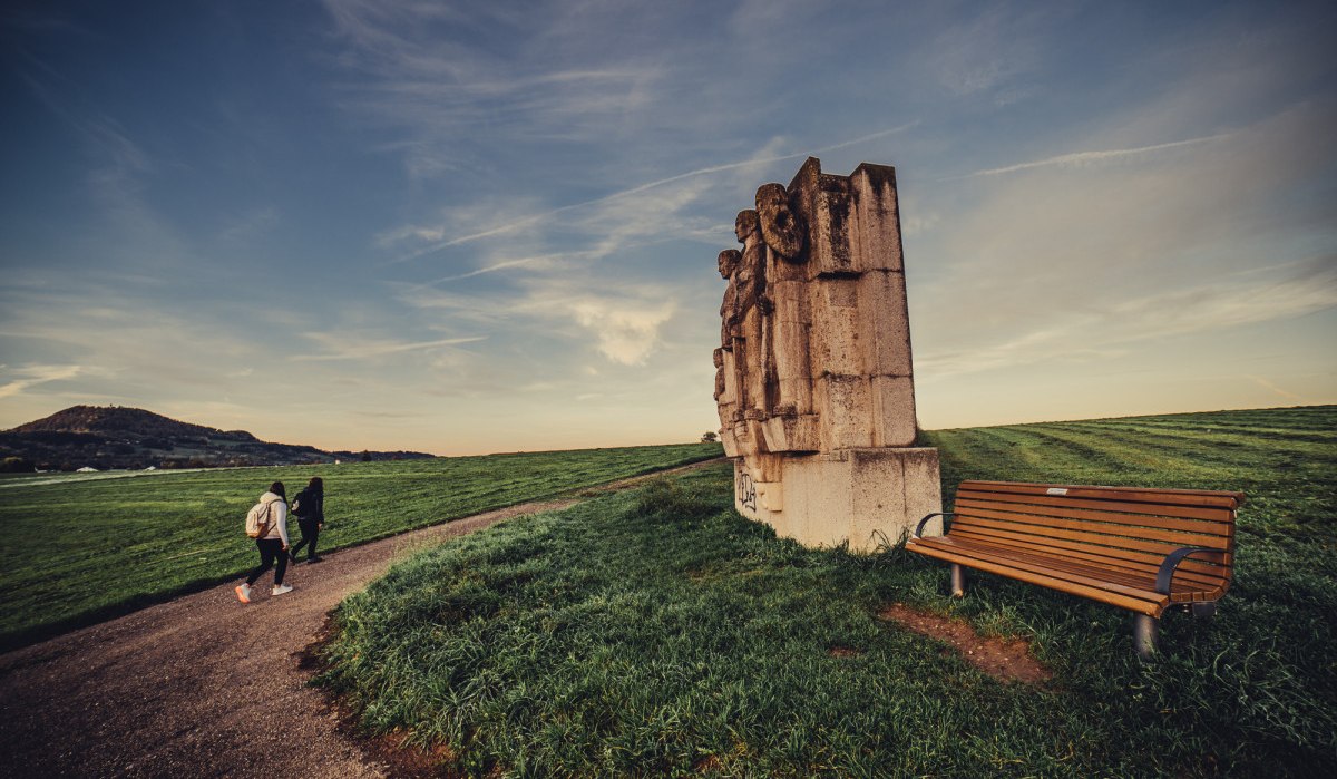 Zwei Personen spazieren auf einem Weg neben einer großen Steinskulptur und einer Bank auf einem grünen Feld unter blauem Himmel., © Landratsamt Ostalbkreis