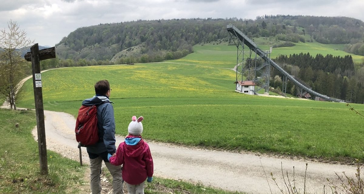 Zwei Personen stehen auf einem Weg und blicken auf eine Sprungschanze in einer gr&uuml;nen Landschaft mit H&uuml;geln und W&auml;ldern., &copy; Touristik und Marketing GmbH