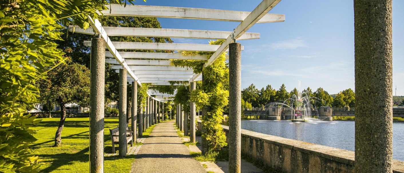 Ein sonniger Parkweg mit Pergola und grünen Pflanzen führt entlang eines Sees mit einem Springbrunnen im Hintergrund., © SMG, Sarah Schmid Ein sonniger Parkweg mit Pergola und grünen Pflanzen führt entlang eines Sees mit einem Springbrunnen im Hintergrund., © SMG, Sarah Schmid