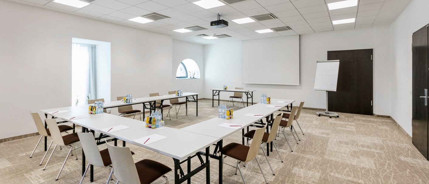 Moderner Konferenzraum mit U-f&ouml;rmiger Tischanordnung, St&uuml;hlen, Flipchart und Projektor. Helle Beleuchtung und neutrale Farben dominieren den Raum., &copy; Arcotel Hotels