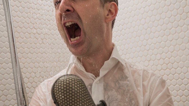 Ein Mann in einem wei&szlig;en Hemd singt leidenschaftlich unter der Dusche mit einem Mikrofon in der Hand. Die Duschwand ist mit runden Fliesen bedeckt., &copy; Scala Kultur Live gGmbH