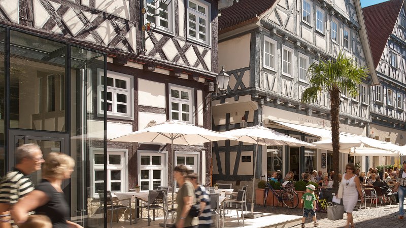 Belebte Stra&szlig;e mit Fachwerkh&auml;usern, Caf&eacute;s und Passanten. Menschen sitzen unter Sonnenschirmen, w&auml;hrend andere vorbeigehen. Palmen schm&uuml;cken die Szene., &copy; Oswald