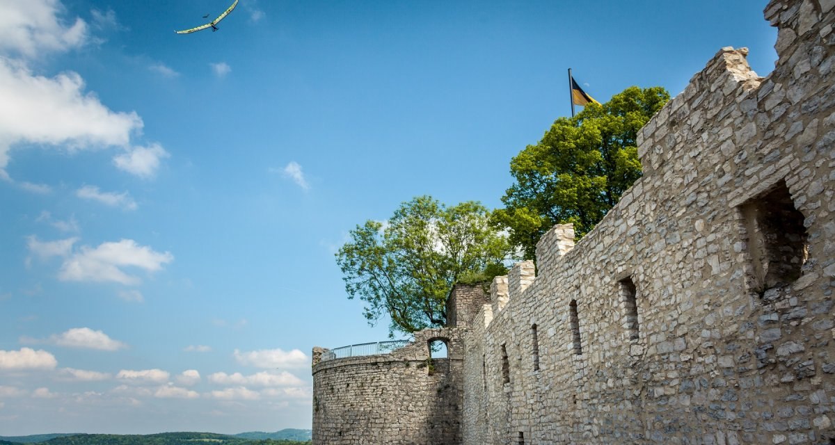Die Burgruine Hohenneuffen mit einer Steinmauer und Bäumen, darüber ein Segelflugzeug am blauen Himmel., © hochgehberge Die Burgruine Hohenneuffen mit einer Steinmauer und Bäumen, darüber ein Segelflugzeug am blauen Himmel., © hochgehberge