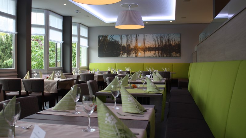 Modernes Restaurant mit gedeckten Tischen, gr&uuml;nen Servietten und gro&szlig;en Fenstern. An der Wand h&auml;ngt ein Bild mit Sonnenuntergangsmotiv., &copy; Hotel am Schlossberg Herrenberg