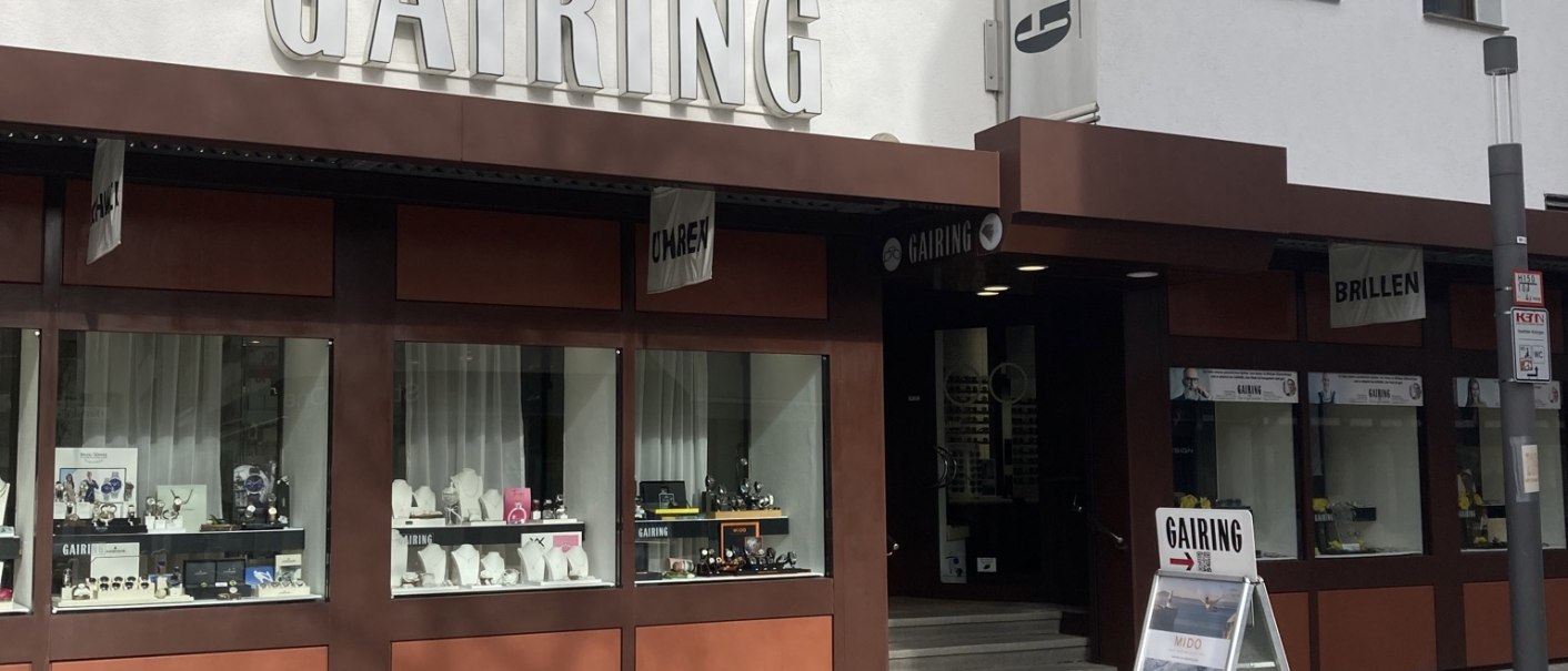 Geschäftsfassade von Gairing mit großen Schaufenstern, die Uhren und Brillen präsentieren. Ein Werbeschild steht vor dem Eingang., © Lena Besemer Geschäftsfassade von Gairing mit großen Schaufenstern, die Uhren und Brillen präsentieren. Ein Werbeschild steht vor dem Eingang., © Lena Besemer