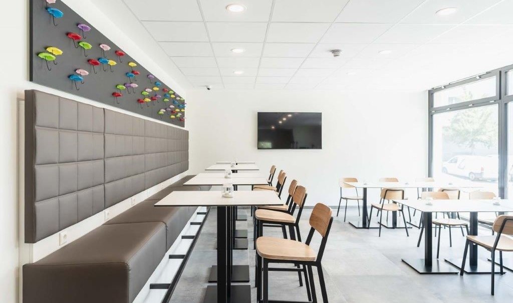 Moderner Frühstücksbereich mit weißen Tischen, Holzstühlen und bunter Regenschirm-Dekoration an der Wand., © a2 HOTELS am Quadrium