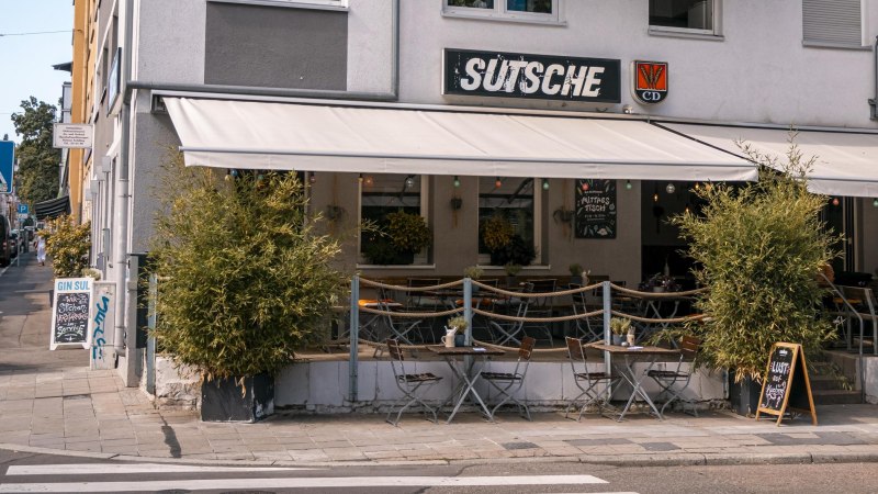Café 'Sutsche' mit Außenbereich, Tischen und Stühlen auf dem Gehweg, umgeben von Pflanzen., © SMG, Sarah Schmid Café 'Sutsche' mit Außenbereich, Tischen und Stühlen auf dem Gehweg, umgeben von Pflanzen., © SMG, Sarah Schmid