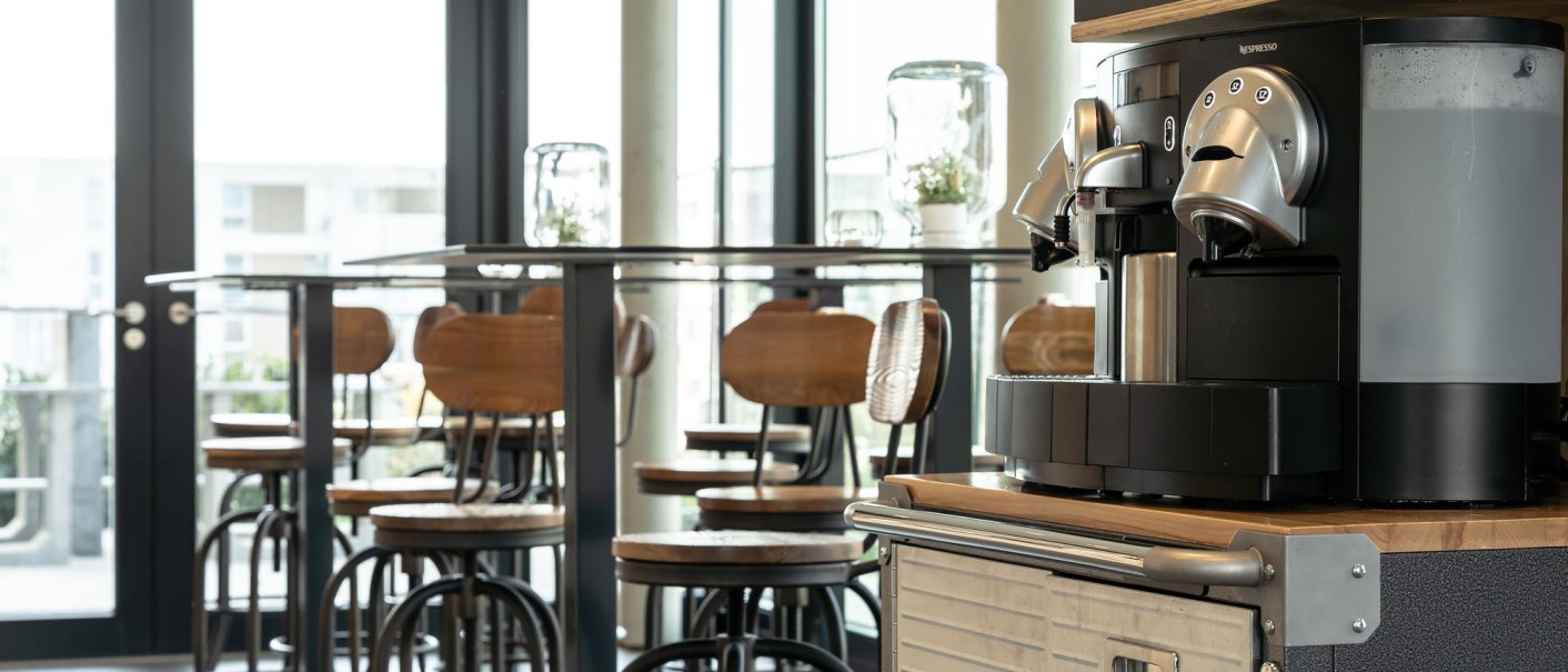 Ein moderner Fr&uuml;hst&uuml;cksbereich mit einer Kaffeemaschine im Vordergrund und hohen Tischen mit St&uuml;hlen im Hintergrund., &copy; a2 HOTELS