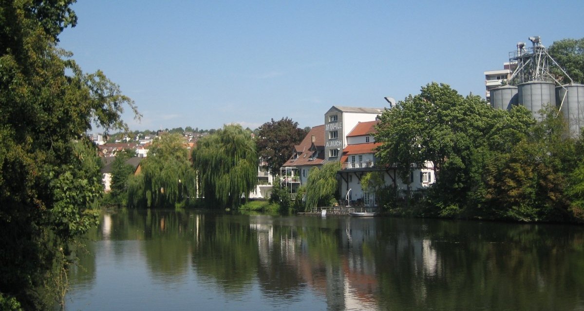 Ein ruhiger Fluss mit Bäumen und Gebäuden am Ufer, darunter eine Mühle mit der Aufschrift 'Mühle'. Der Himmel ist klar und blau., © Land der 1000 Hügel - Kraichgau-Stromberg Ein ruhiger Fluss mit Bäumen und Gebäuden am Ufer, darunter eine Mühle mit der Aufschrift 'Mühle'. Der Himmel ist klar und blau., © Land der 1000 Hügel - Kraichgau-Stromberg