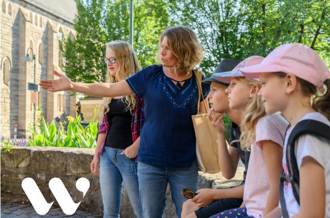 Eine Frau zeigt einer Gruppe von Kindern w&auml;hrend einer Stadtf&uuml;hrung etwas Interessantes. Im Hintergrund ist ein historisches Geb&auml;ude und gr&uuml;ne B&auml;ume zu sehen., &copy; Stadt Winnenden