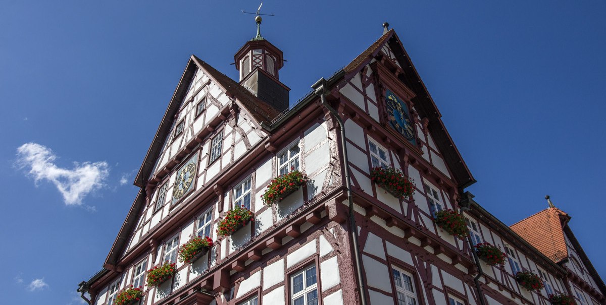 Das historische Fachwerk-Rathaus von Bad Urach mit blühenden Blumenkästen und einer Uhr, vor einem klaren blauen Himmel., © Bad Urach Tourismus