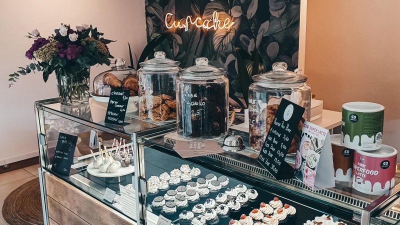 Ein Caf&eacute; mit einer Glasvitrine voller Cupcakes und Kekse. Ein Neon-Schild mit 'Cupcake' leuchtet im Hintergrund. Blumen schm&uuml;cken den Tresen., &copy; Cupcakes & Bagels, Stuttgart