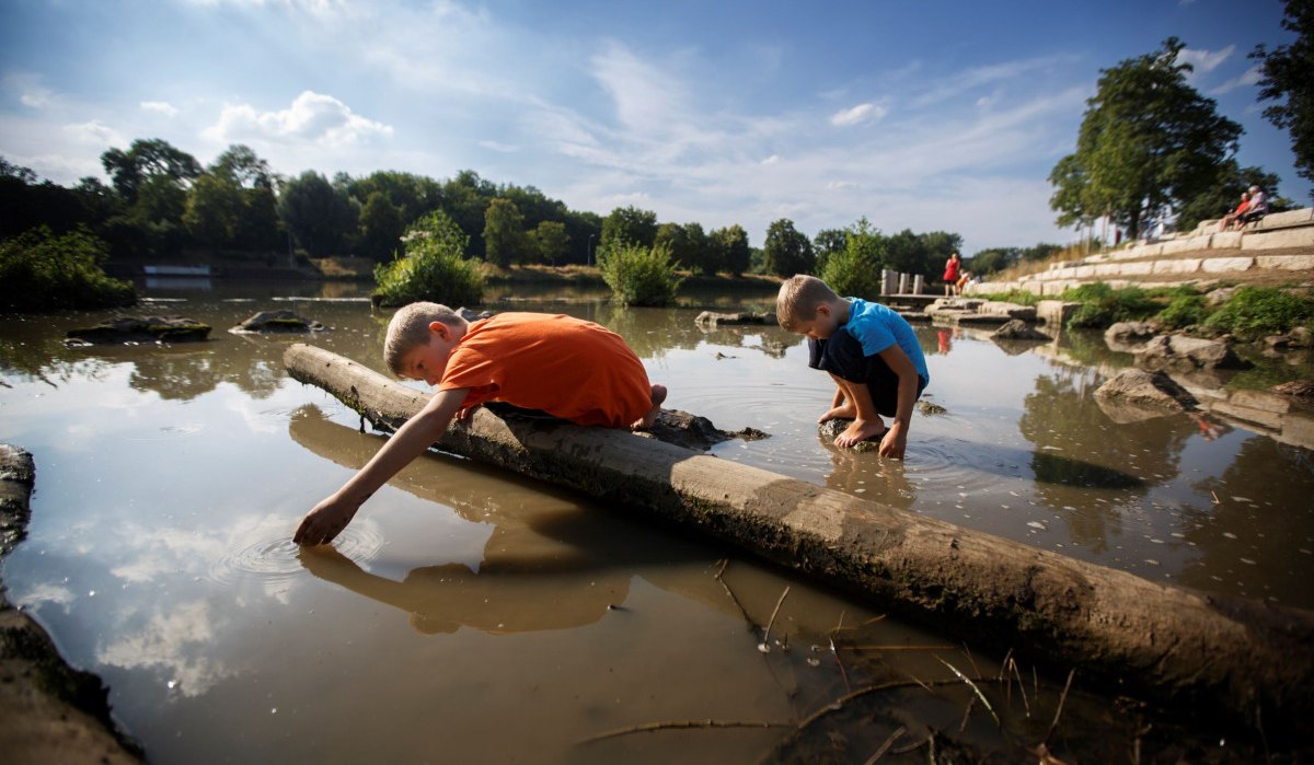 Zwei Kinder spielen am Ufer eines Flusses. Eines liegt auf einem Baumstamm, das andere kniet im Wasser. Im Hintergrund sind Bäume und ein sonniger Himmel., © Stoppel Zwei Kinder spielen am Ufer eines Flusses. Eines liegt auf einem Baumstamm, das andere kniet im Wasser. Im Hintergrund sind Bäume und ein sonniger Himmel., © Stoppel