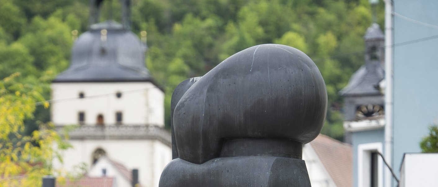 Moderne Skulptur in einer städtischen Umgebung, im Hintergrund ein Kirchturm und grüne Bäume. Die Skulptur ist abstrakt und dunkel gefärbt., © Archiv Würth