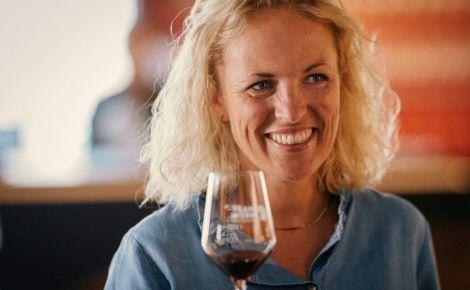 Eine Frau mit blonden Haaren l&auml;chelt und h&auml;lt ein Weinglas. Sie tr&auml;gt ein blaues Hemd. Der Hintergrund ist unscharf., &copy; Tag des offenen Glases; Foto: Peter Bender