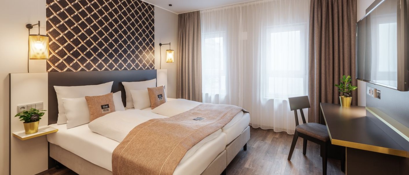 Modernes Hotelzimmer mit Doppelbett, dekorativer Wand, Schreibtisch und Pflanzen. Helle Vorhänge und warme Beleuchtung schaffen eine gemütliche Atmosphäre., © Novum Hospitality Modernes Hotelzimmer mit Doppelbett, dekorativer Wand, Schreibtisch und Pflanzen. Helle Vorhänge und warme Beleuchtung schaffen eine gemütliche Atmosphäre., © Novum Hospitality