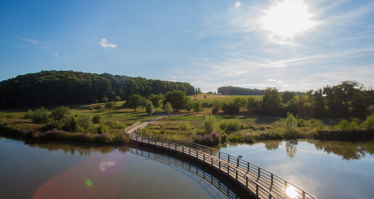 Holzsteg &uuml;ber einen Fluss in einer gr&uuml;nen Landschaft mit B&auml;umen und Wiesen, unter einem strahlend blauen Himmel mit Sonne., &copy; Tourismus und Events Ludwigsburg