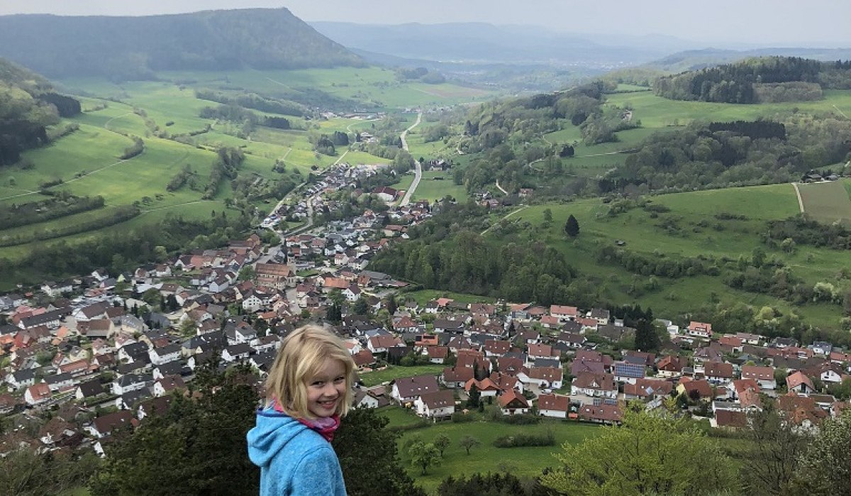 Ein Kind in blauer Jacke steht auf einem Felsen und blickt auf ein Dorf in einer grünen Hügellandschaft., © Touristik und Marketing GmbH
