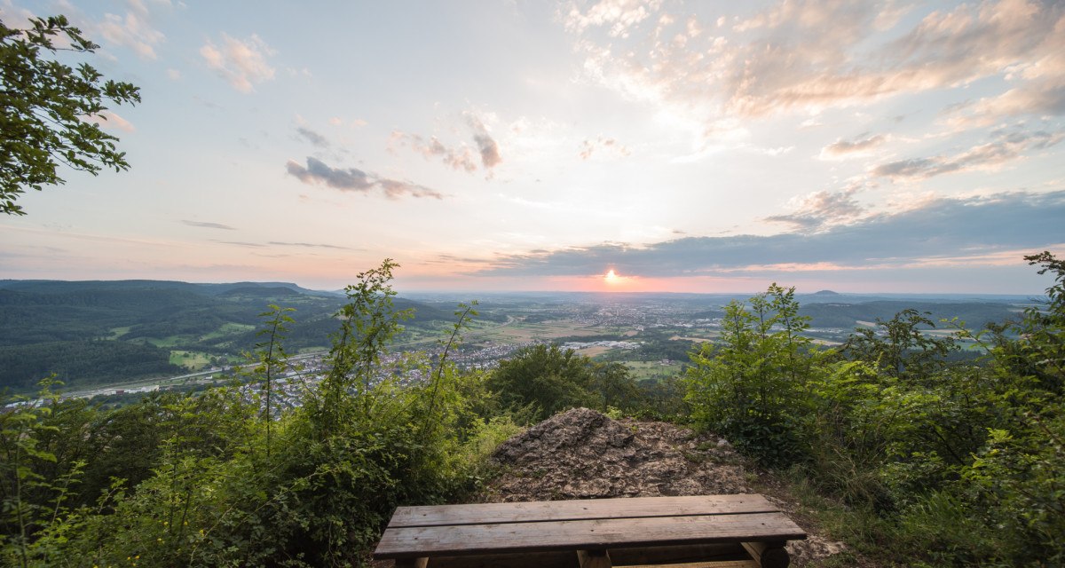 Aussicht vom Hohenstein: Eine Holzbank steht vor einer weiten Landschaft mit Wäldern und Feldern, während die Sonne am Horizont untergeht., © Landkreis Göppingen Aussicht vom Hohenstein: Eine Holzbank steht vor einer weiten Landschaft mit Wäldern und Feldern, während die Sonne am Horizont untergeht., © Landkreis Göppingen